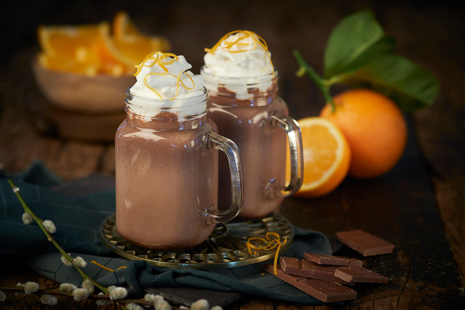 Hjemmelaget kakao med appelsin og pisket krem servert i to glasskrus på rustikk mørkt trebord. Halv og hel appelsin med stilk i bakgrunnen. Gåsunger på stilk, og biter med sjokolade i forgrunnen.