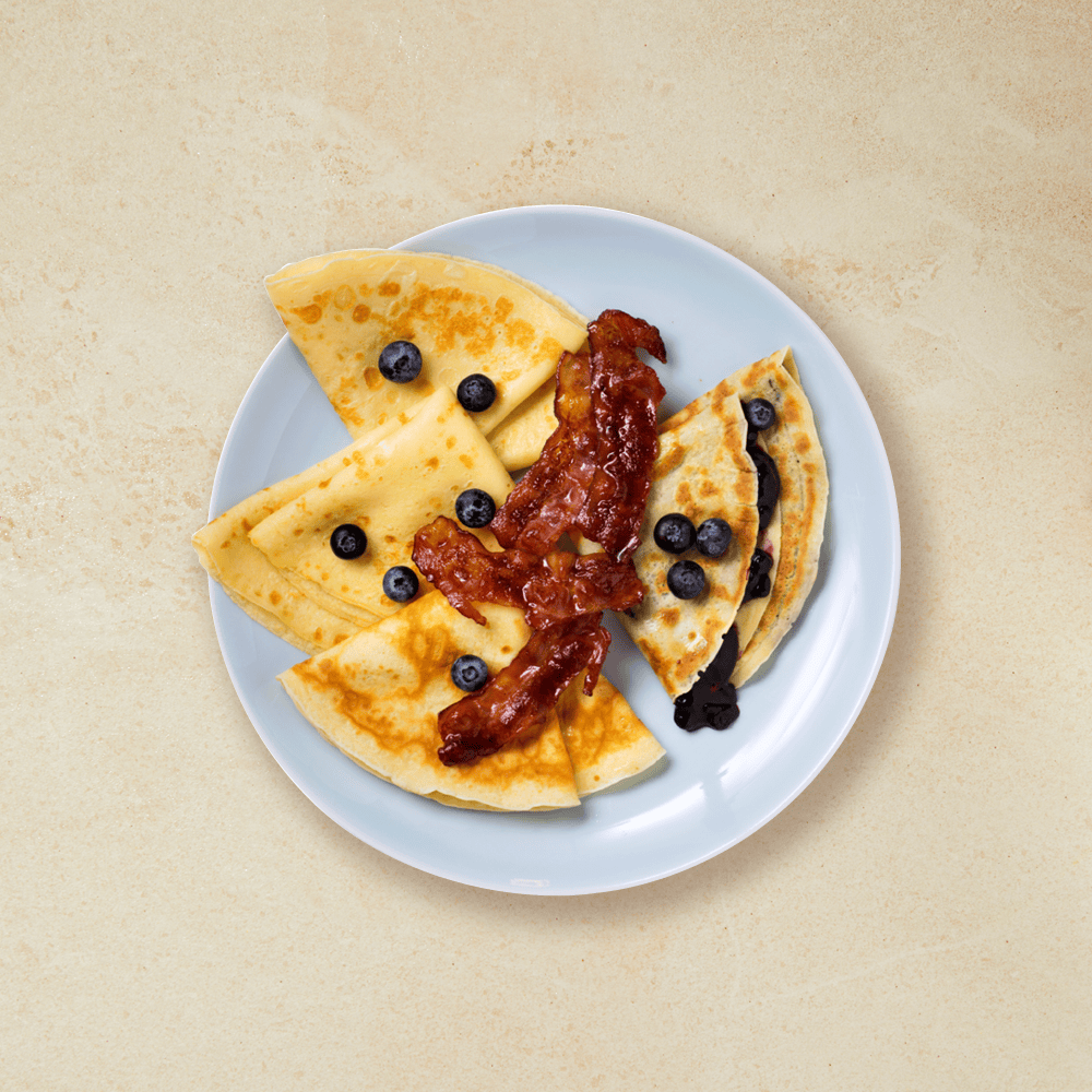 Fire pannekaker fylt med blåbærsyltetøy, pyntet med blåbær og bacon på en lyseblå tallerken.