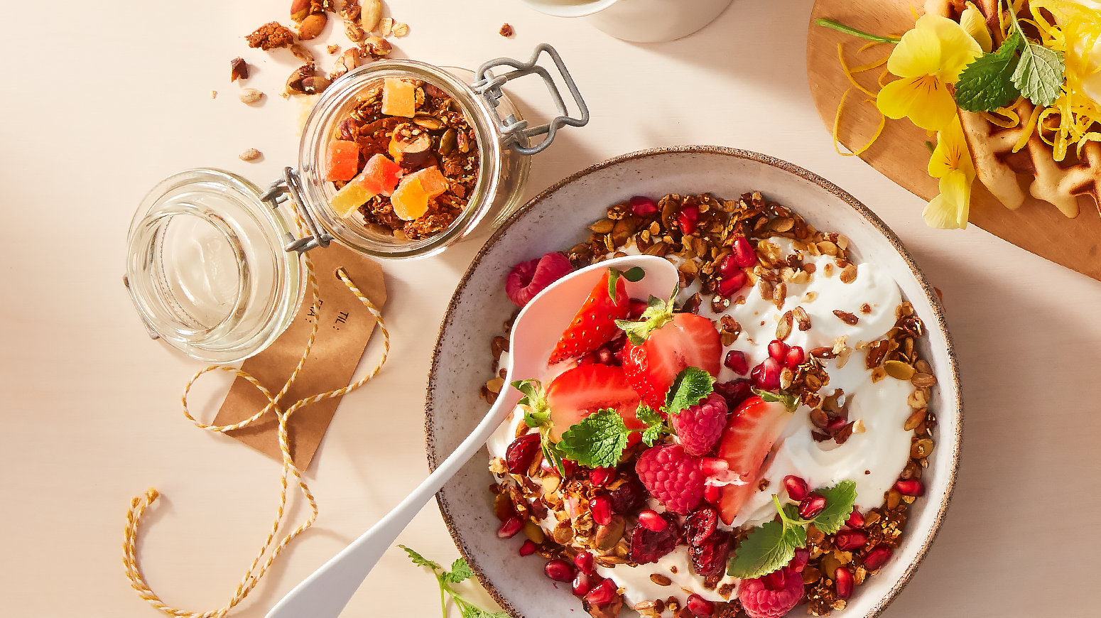 Hjemmelaget granola med tørket frukt i syltetøglass ved siden av en serveringsskål med yoghurt, hjemmelaget granola, ferske bringebær og jordbær, og friske urter. Skålen står på lyst sommerlig bord.