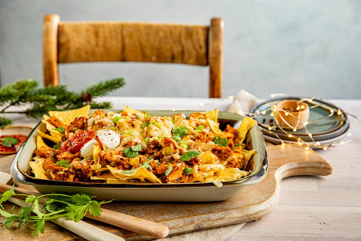 Nachos med rester av kalkunkjøtt, gylden smeltet ost, vårløk, rømme og salsa, servert i ildfast form på lys trefjæl. Hint av jul i bakgrunnen.