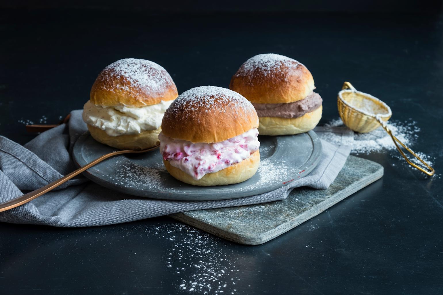 Fastelavnsboller med 3 typer kremfyll servert på grå tallerken, med grått kjøkkenhåndkle under. Antikk sikt med melis ved siden av.