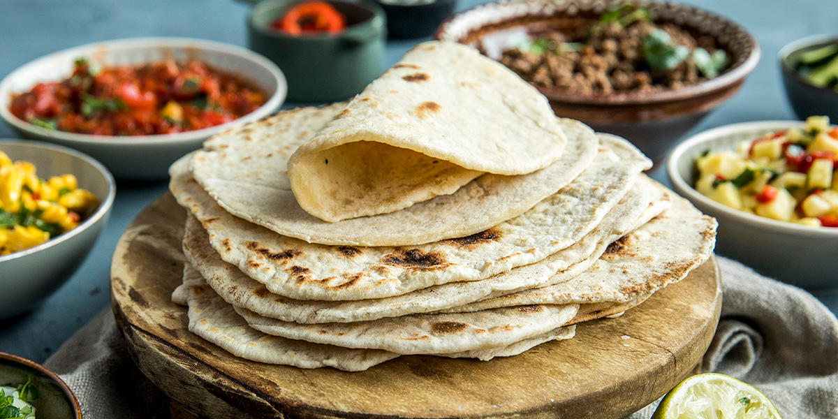 Hjemmelagde tortillalefser til taco, stablet på trefjøl. I bakgrunnen står skåler med tacoingredienser som stekt kjøttdeig, tomatsalsa og mangosalsa.