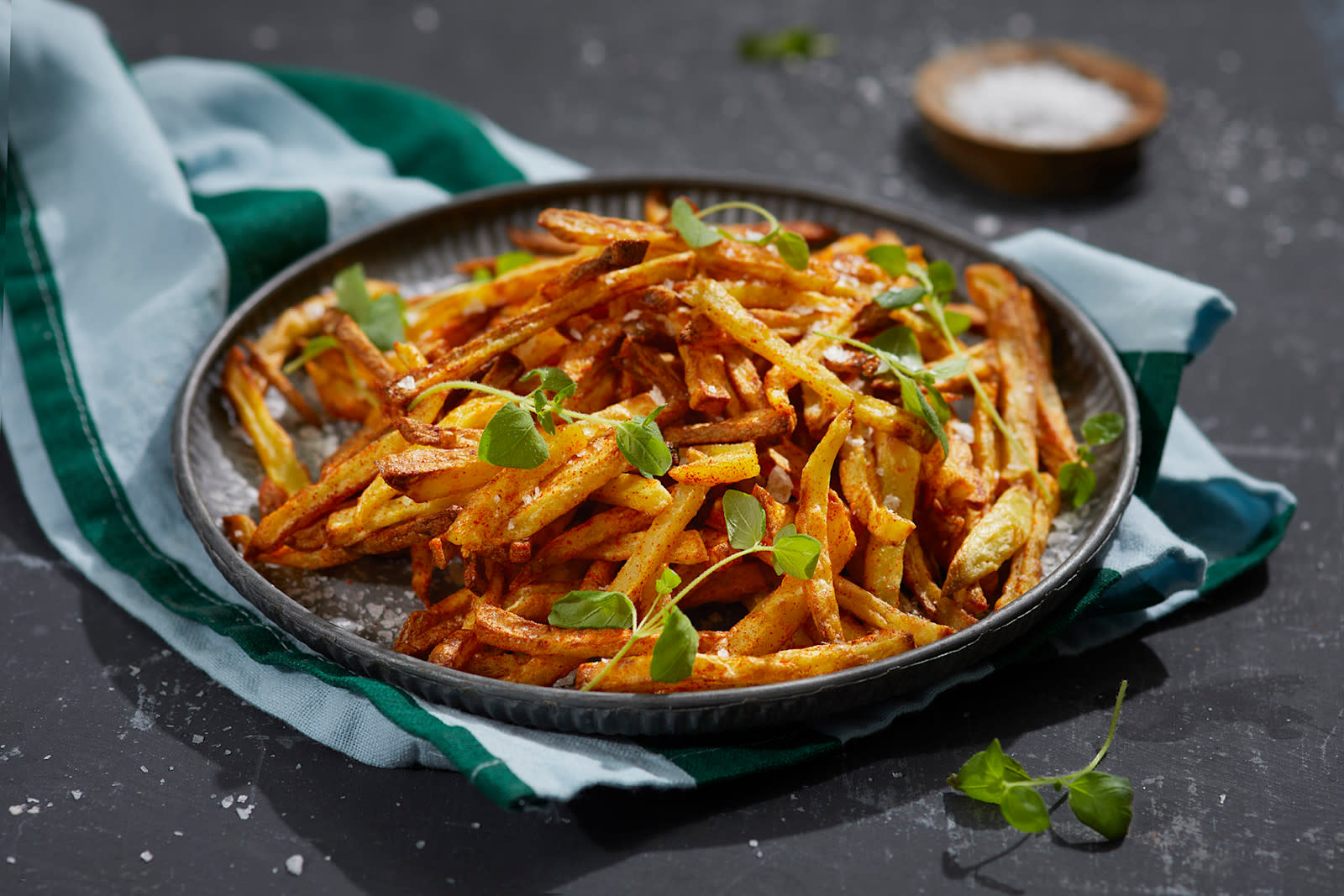 Skinny fries laget i airfryer servert på mørkegrått rundt fat med grovt salt og grønne urter over. Liten skål med grovt salt i bakgrunnen.