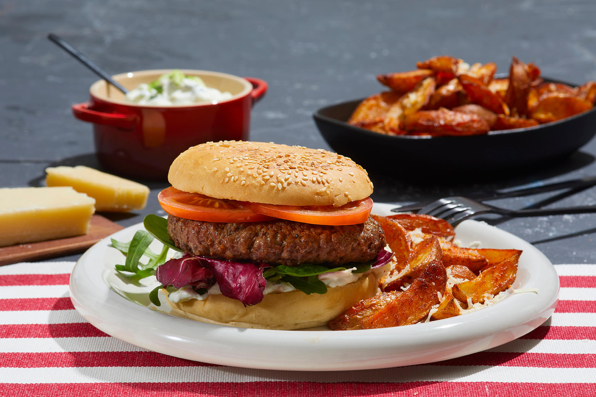 Big Beefy burger med hjemmelaget tzatziki og potetbåter med parmesan, servert på hvit tallerken på hvit og rødstripet duk. Skåler med tzatziki og potetbåter i bakgrunnen.