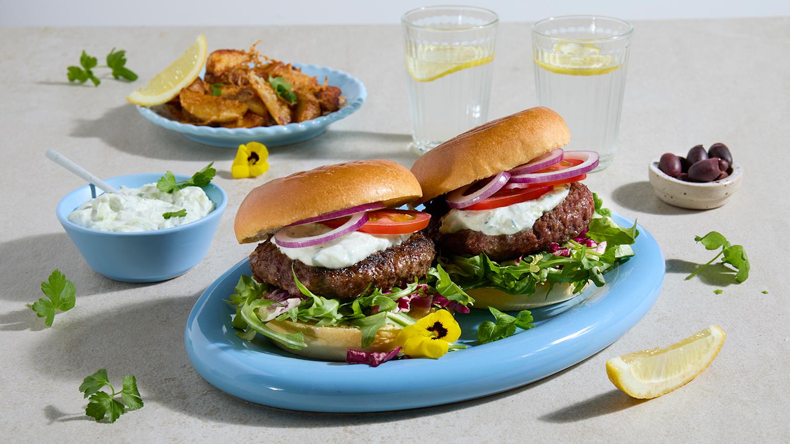 Big Beefy burger med hjemmelaget tzatziki og parmesanpoteter servert på lyseblå asjett. Skåler med tzatziki og parmesanpoteter i bakgrunnen.