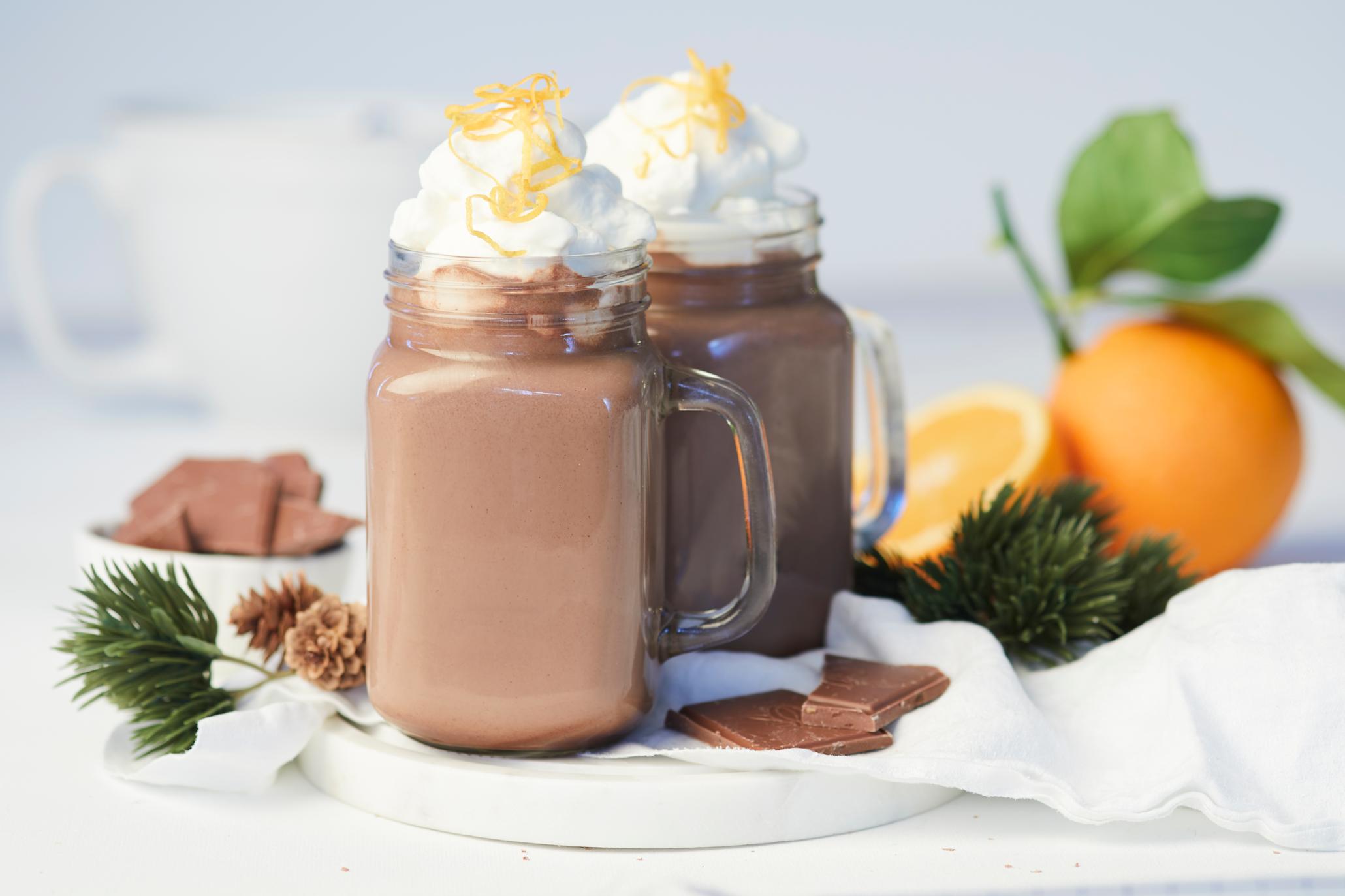 Hjemmelaget kakao med appelsin og pisket krem servert i glasskrus på hvitt rundt marmorfat med biter av sjokolade og granbar rundt. Hel appelsin med blad, og halv appelsin i bakgrunnen.