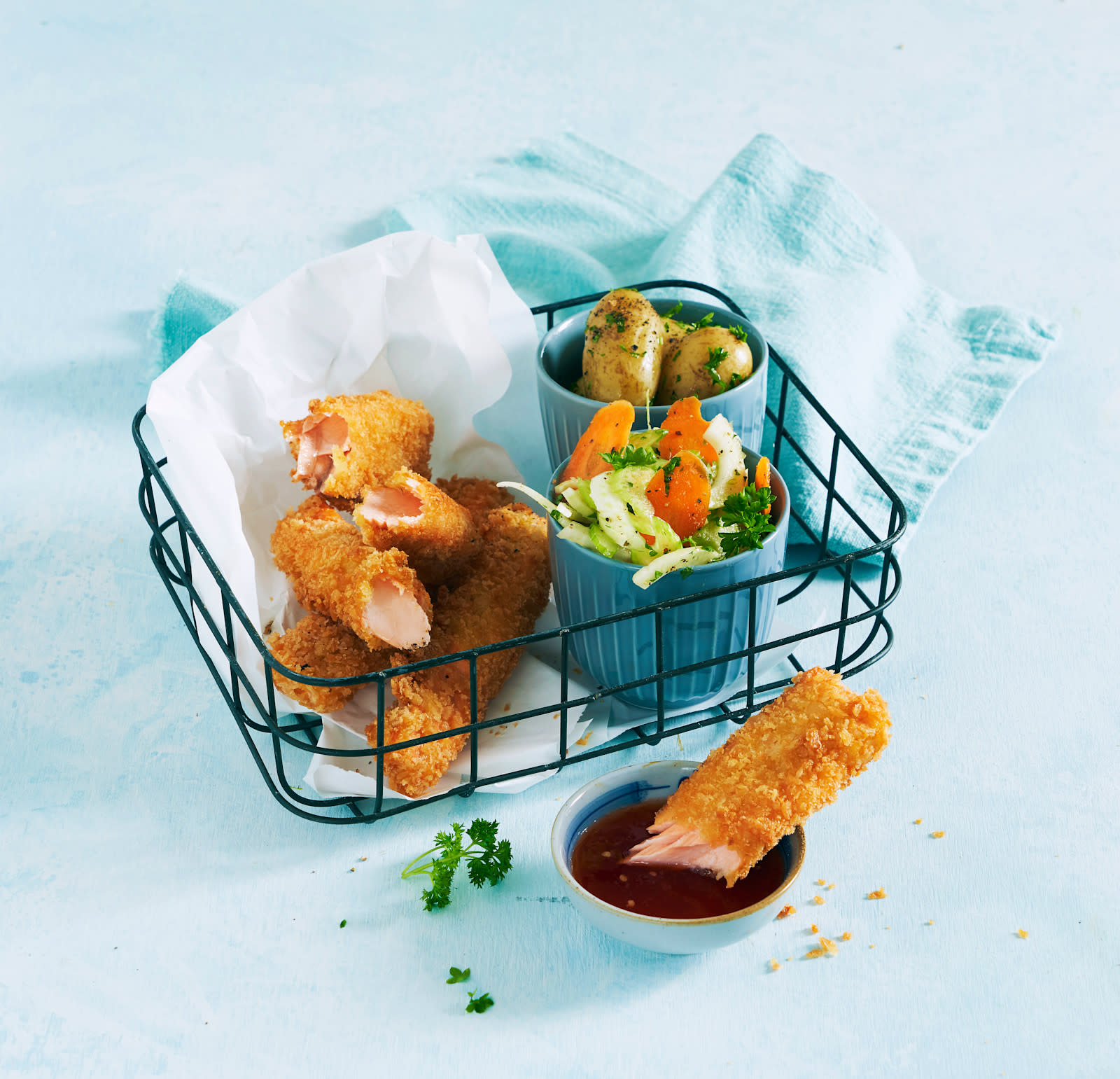 Hjemmelagde fiskepinner av laks med frisk råkostsalat servert på hvitt bakepapir i metallkurv med kokte amandinepoteter og rød dip ved siden av. Blå bakgrunn.