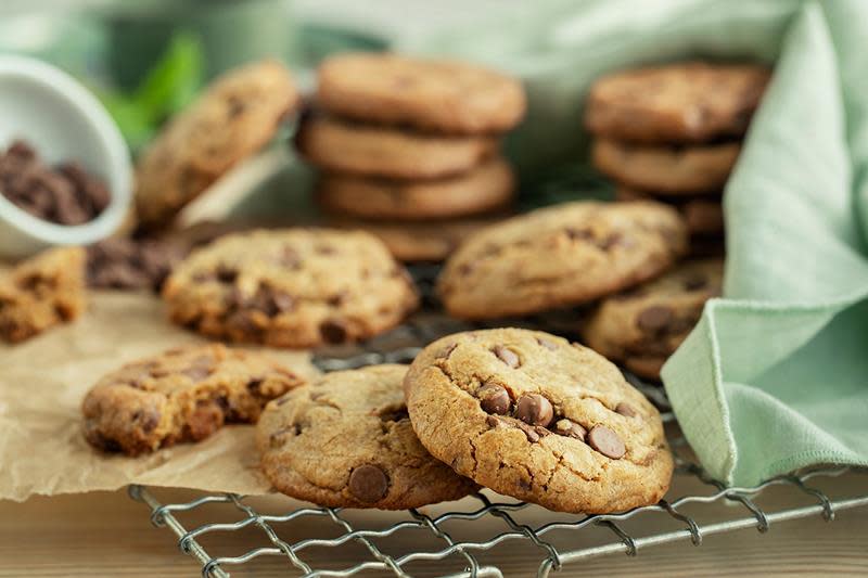 Store og myke sjokoladekjeks med sjokoladedråper servert på avkjølingsrist i metall, med bakepapir over. Lyst grønt kjøkkenhåndkle lagt halvveis over kjeksene, og en liten krukke sjokoladedråper ved siden av.