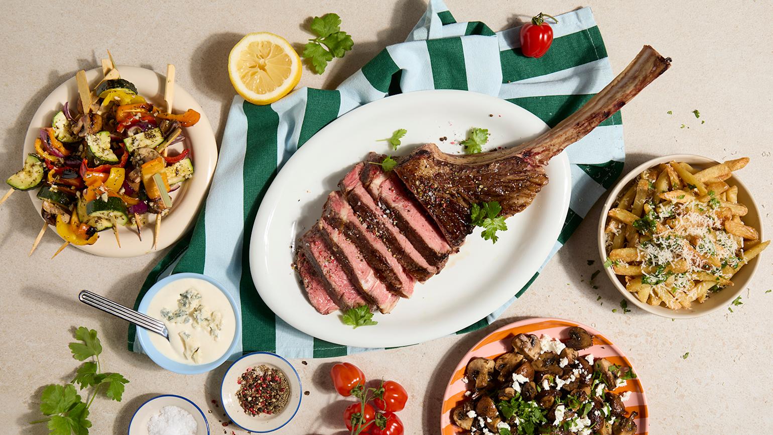 Reverse sear tomahawk servert i skiver på oval hvit asjett med skåler med tilbehør rundt.