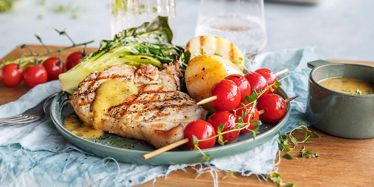 Sommerkoteletter med sennepssaus og grillet hjertesalat, servert på grønn tallerken med nypoteter og cherrytomater. Tallerkenen står på en trefjøl, med en liten skål sennepssaus ved siden av.