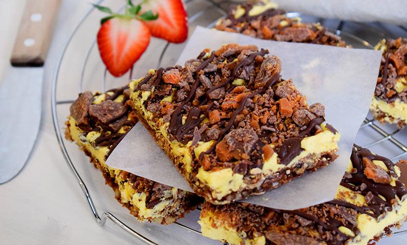 Daimkake i langpanne med nøttebunn og gul krem, skåret i stykker, og servert på kjølerist i metall. Stykkene er toppet med smeltet sjokolade og hakket Daim. Ferske jordbær ved siden av.