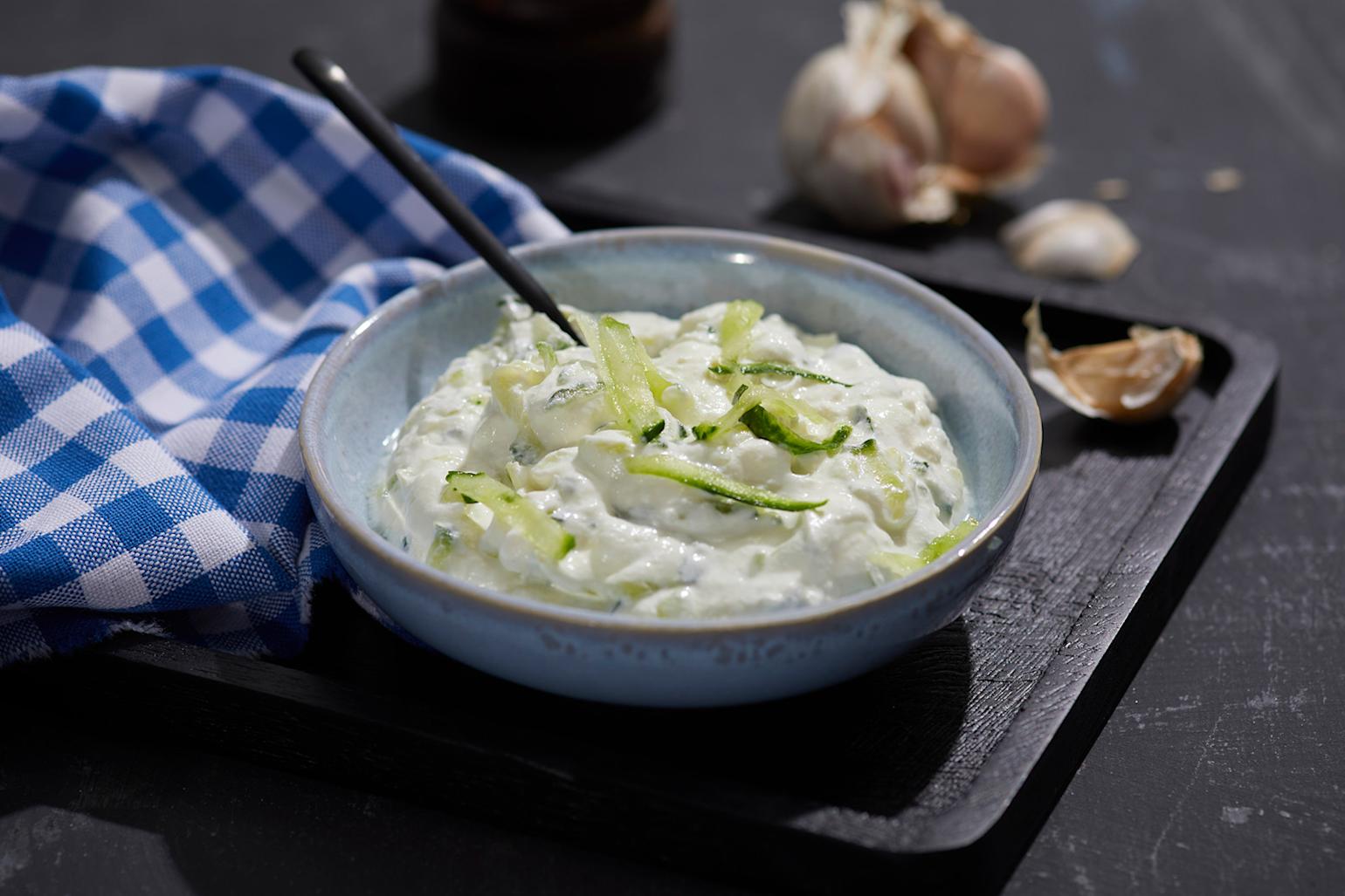 Hjemmelaget tzatziki med finrevet agurk på toppen, servert i blågrå keramikkskål på svart serveringsbrett. Blårutet kjøkkenhåndkle på siden, og hvitløksbåter i bakgrunnen.
