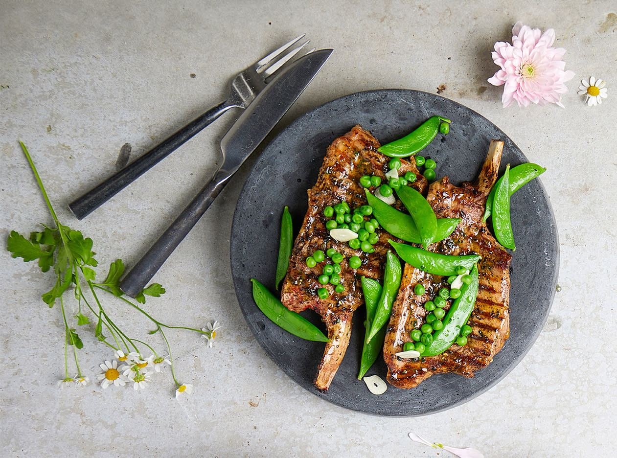 Marinerte koteletter med bein, servert på mørkegrå tallerken med sukkererter, erter og skivet hvitløk. Tallerkenen står på en lys steinflate med forskjellige blomster.