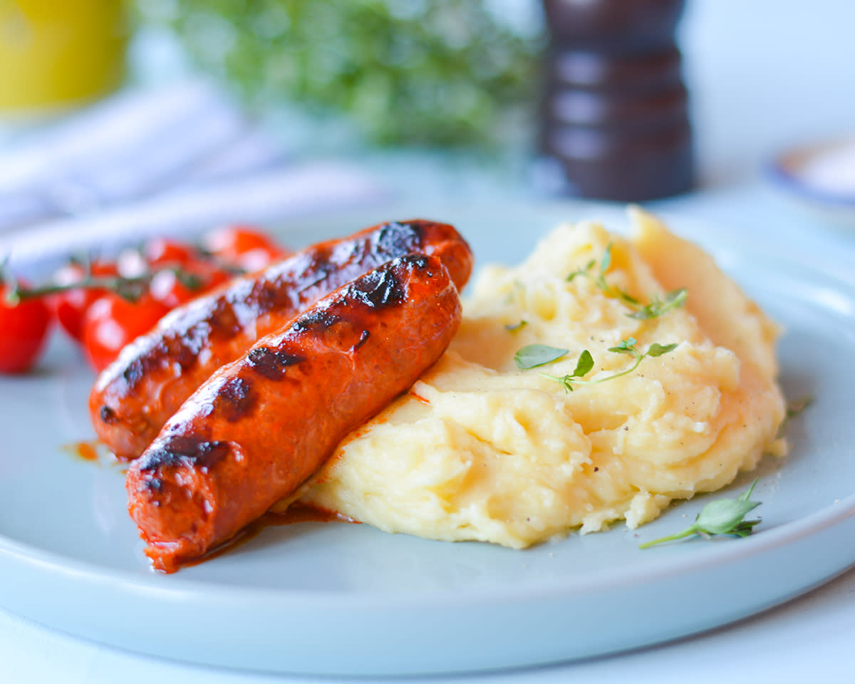 Fransk potetmos med stekt pølse på blå tallerken med tomater i bakgrunnen.