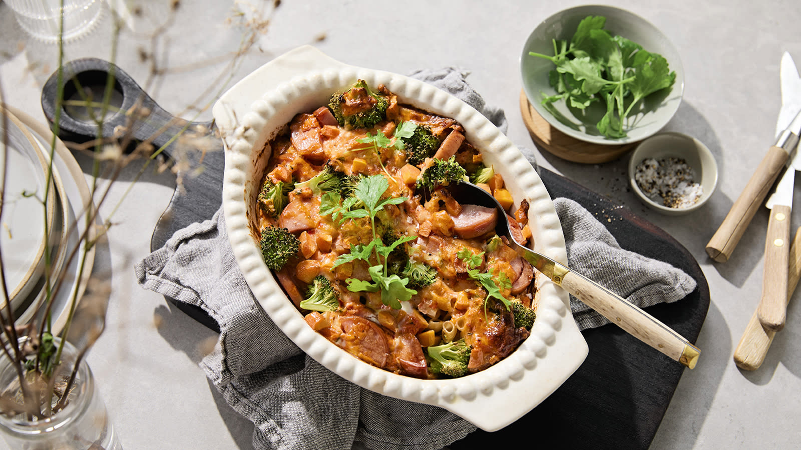 Pølsegrateng med pasta og brokkoli servert i hvit ildfast form på mørk trefjøl. Skåler med krydder og ruccolasalat ved siden av.