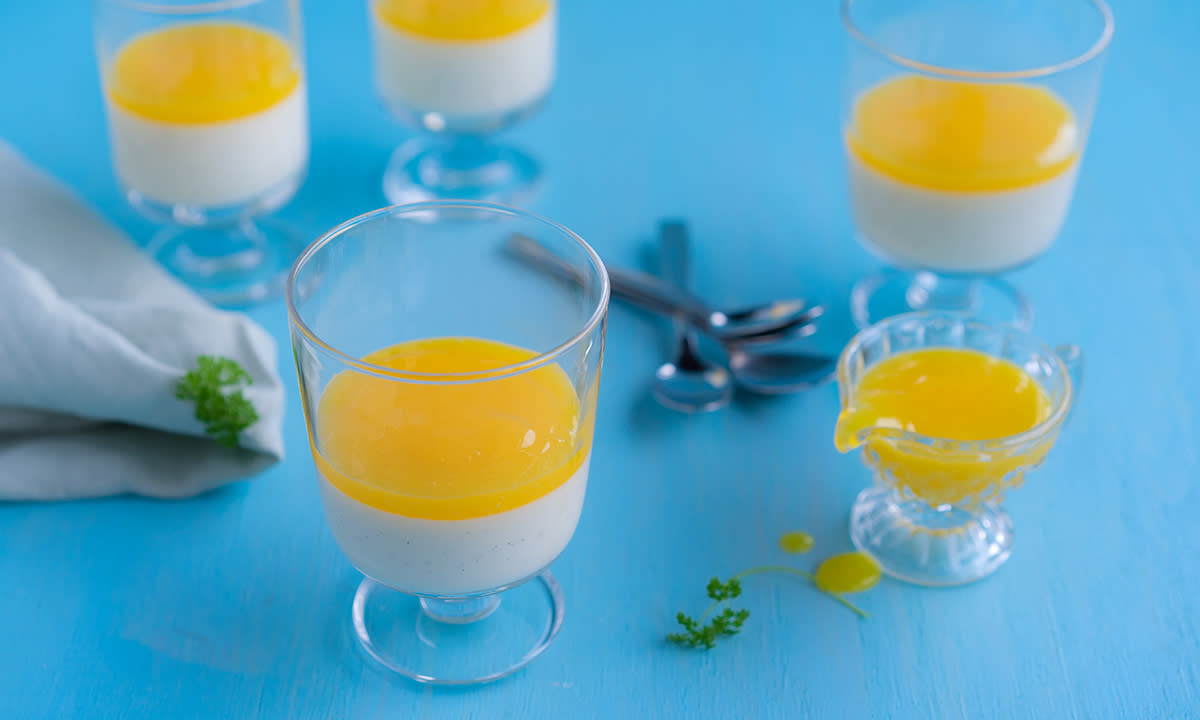 Panna cotta med mangosaus servert i lave stettglass på blå bakgrunn med teskjeer, linserviett og en liten mugge med mangosaus.