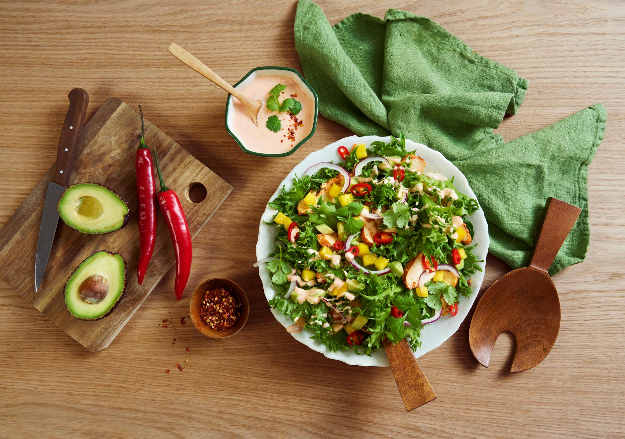 Crispisalat med kylling, mango og sunn dressing servert i hvit skål på lyst trebord. Skjærefjøl med avokado og chilli ved siden av. 
