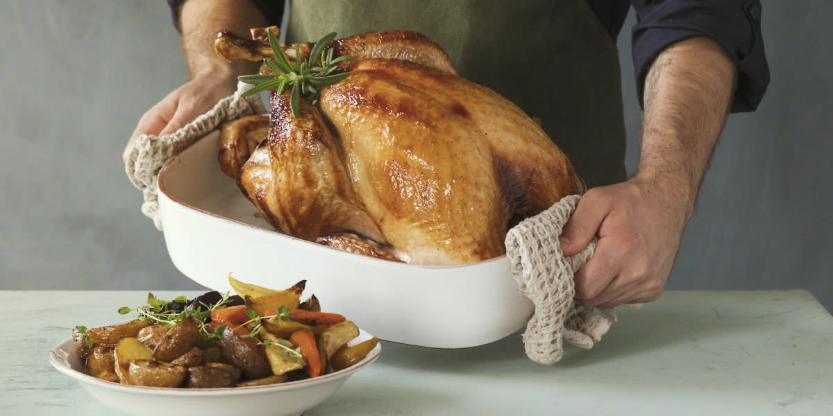 Glasert helstekt kalkun med eplesjy servert i hvit ildfast form av to hender som holder formen frem over et bord. Rosa mønstret skål med ovnsbakte rotgrønnsaker ved siden av.