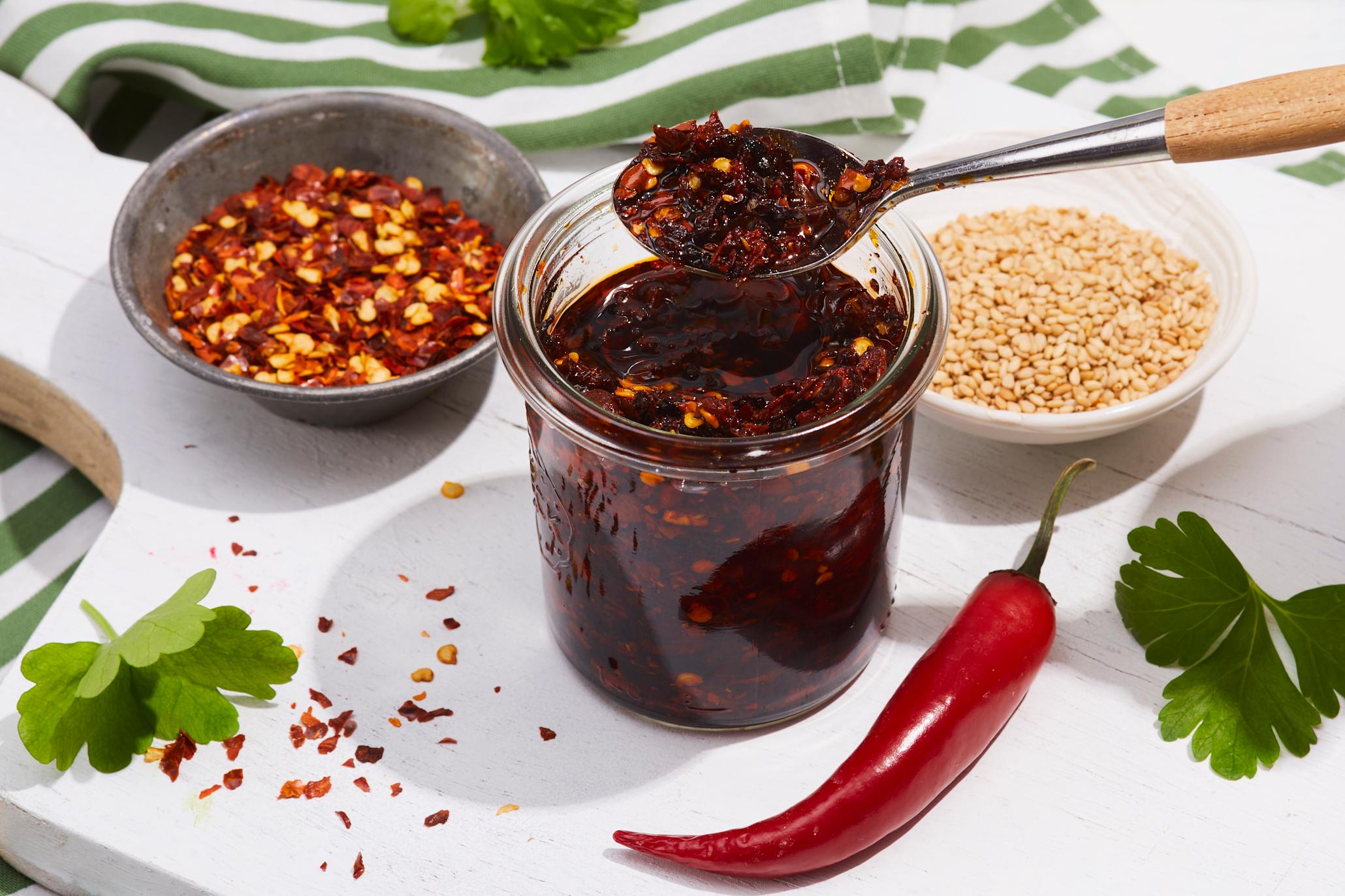 Hjemmelaget chili crisp på lite glass, servert på hvit trefjøl med små skåler chiliflak og sesamfrø ved siden av. Frisk hel chili, og grønne urter rundt.