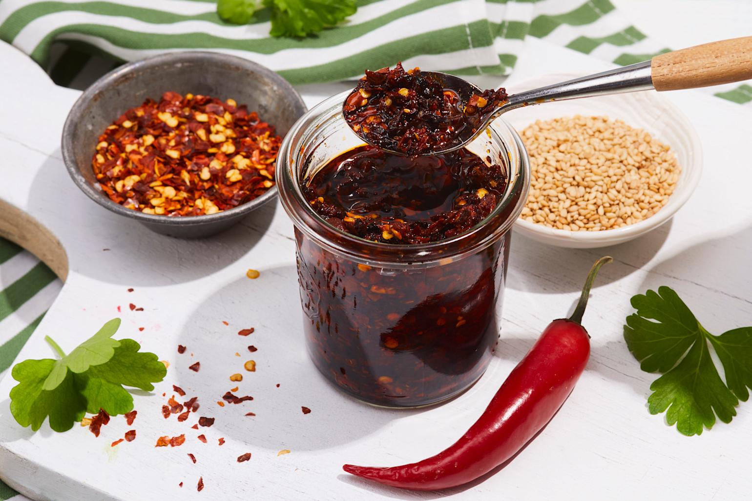 Hjemmelaget chili crisp på lite glass, servert på hvit trefjøl med små skåler chiliflak og sesamfrø ved siden av. Frisk hel chili, og grønne urter rundt.