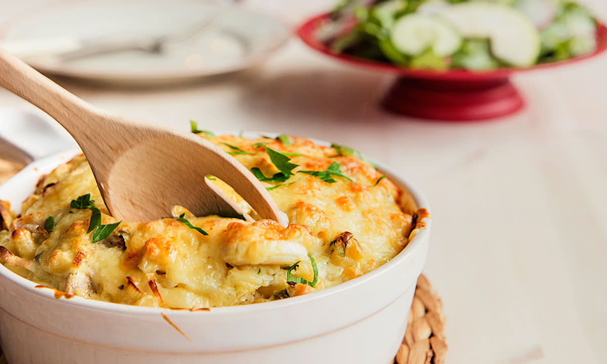 Pølsegrateng med julepølse servert i rund hvit ildfast form, med en tresleiv stukket ned i gratengen. Frisk grønn salat i bakgrunnen.