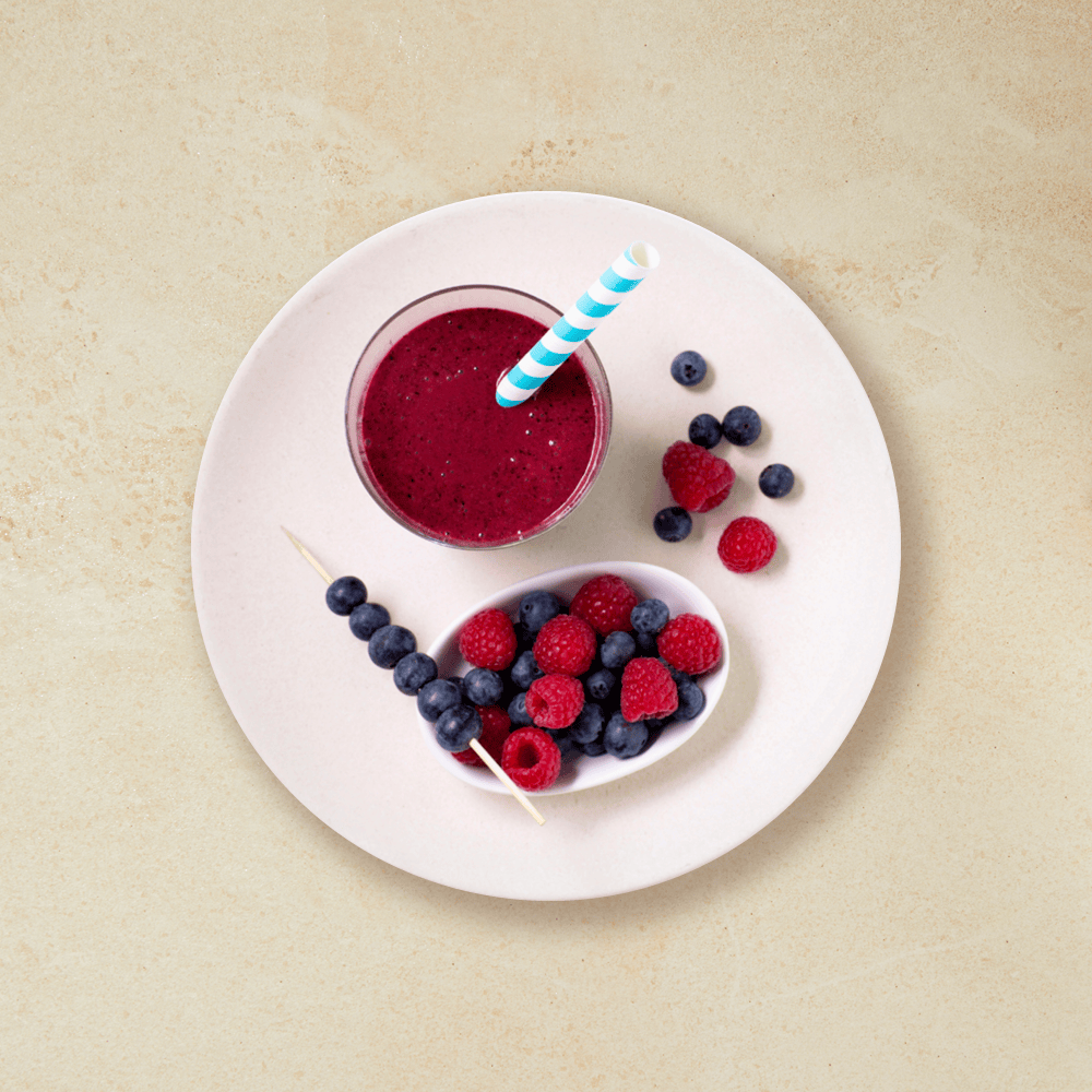 Smoothie med bær servert i smoothieglass på en rosa tallerken, med en skål blåbær og bringebær, og seks blåbær tredd på en tannpirker.