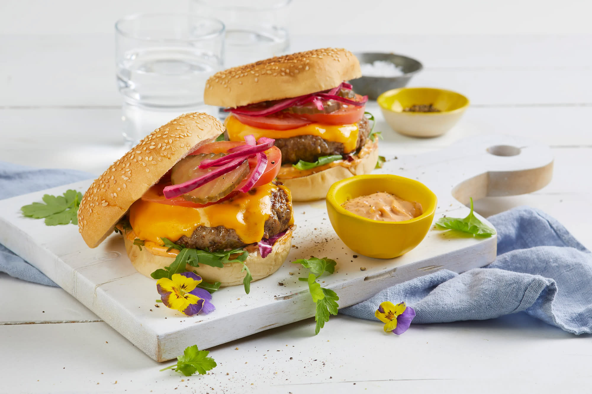 Beefy burger med cheddar og hjemmelaget syltet rødløk i burgerbrød med sylteagurk, tomat og salat, servert på hvit trefjøl på hvitt trebord. Liten gul skål med burgerdressing på siden.