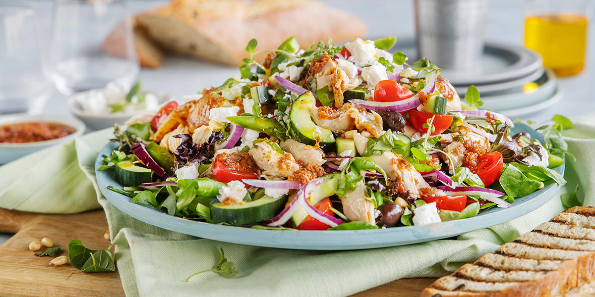 Gresk kyllingsalat med fetaost, oliven og hjemmelaget tomatdressing servert med agurk og urter på lyseblått fat på dekket middagsbord, med ristet brød ved siden av.