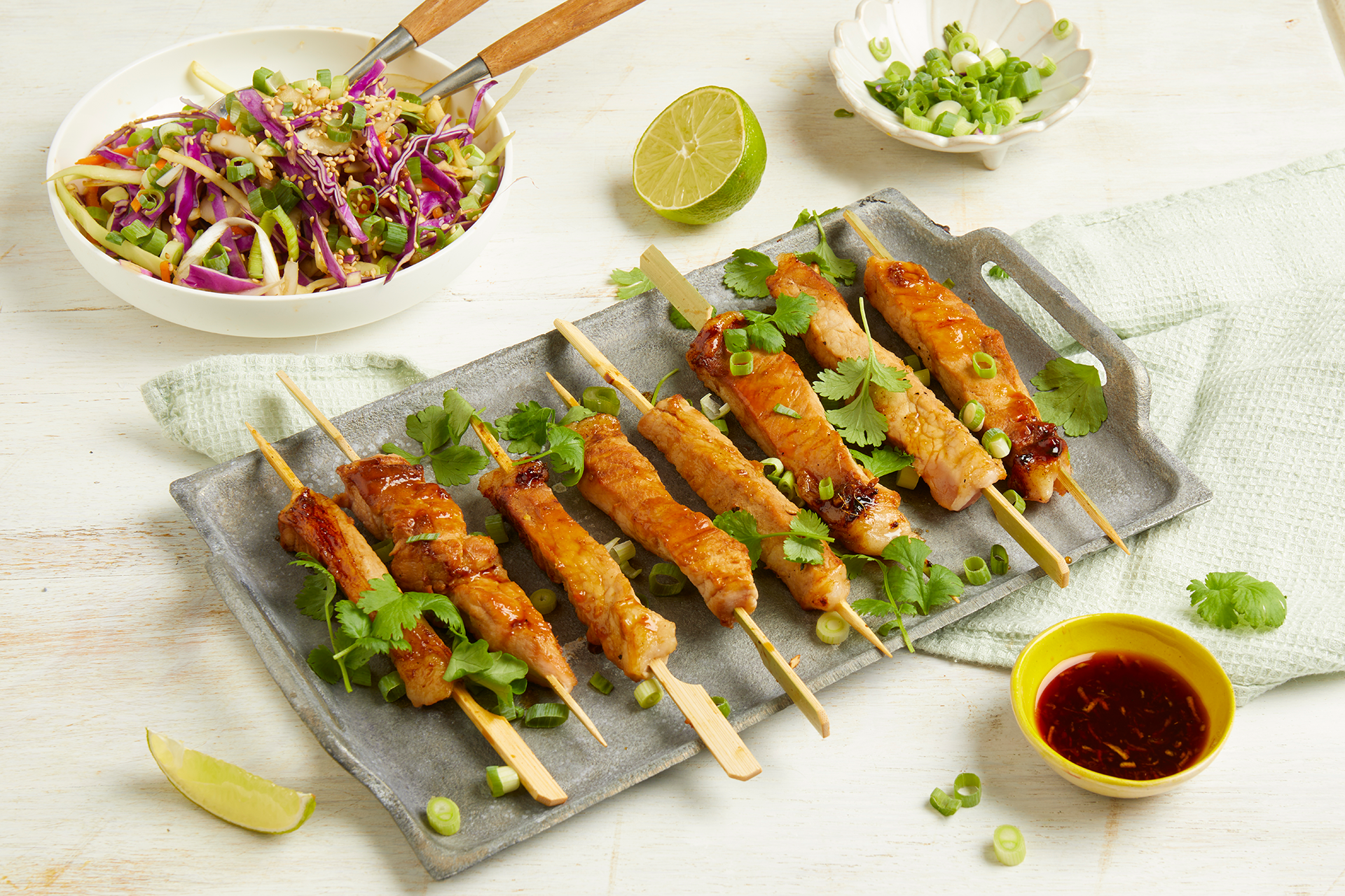 Asiatiske grillspyd av sommerkoteletter med frisk råkostsalat servert på grått fåt på hvitt trebord. Lite gul skål med asiatisk dressing i forgrunnen, og to skåler med asiatisk råkostsalat og finhakket vårløk i bakgrunnen.