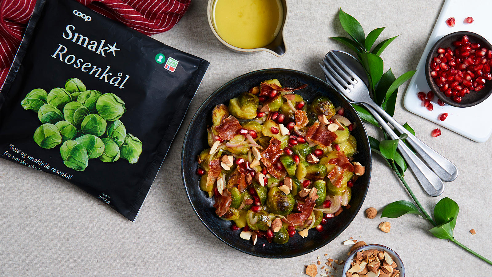 Lun rosenkålsalat med spekeskinke og sennepsvinaigrette servert i svart skål med granateplekjerner og skivede mandler. Liten skål med granateplekjerner, og lite sausenebb med dressing på siden.