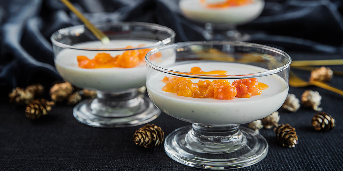 Yoghurt-panna cotta med multer servert i lave dessertglass med stett, på bord med mørk duk og gullfargede små kongler. Gullfargede teskjeer i bakgrunnen.