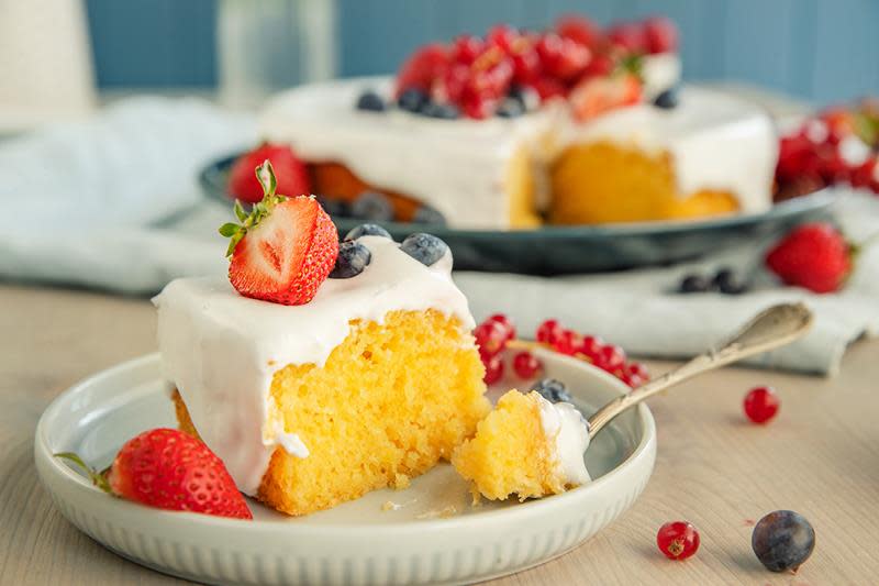 Saftig sitronkake med yoghurt, pyntet med melisglasur, friske jordbær, rips og blåbær. Kaken er servert i stykke på liten asjett foran et fat med resten av kaken.
