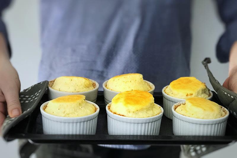 Ostesufflé laget i seks små hvite ildfaste former. Formene er plassert på svart stekebrett som holdes frem av to hender med grytekluter.