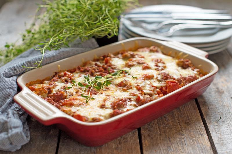 Lasagne i rød ildfast form, toppet med smeltet ost og fersk timian, servert på rustikk trebord. Grå linserviett, stabel med tallerkner og fersk timianplante ved siden av.