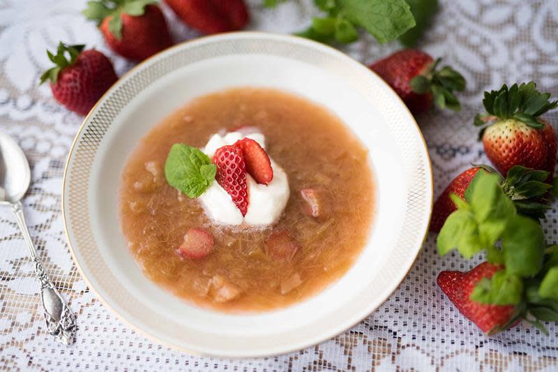 Krydret rabarbrasuppe servert i hvit skål med gullkant, toppet med pisket krem og ferske jordbær. Hvit blondeduk, sølvskje og ferske jordbær i bakgrunnen.
