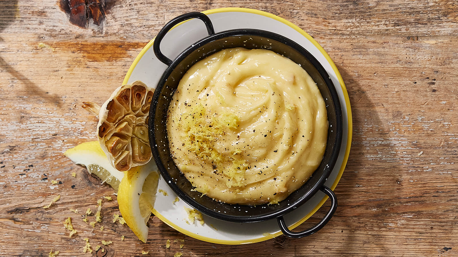 Aioli med bakt hvitløk servert i liten svart skål med revet sitronskall på toppen og en halv bakt hvitløk på siden.