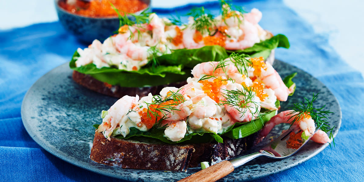Brødskiver med klassisk skagenrøre, servert med salat, rogn og fersk dill på blå tallerken på blå duk. Blå skål med rogn i bakgrunnen.