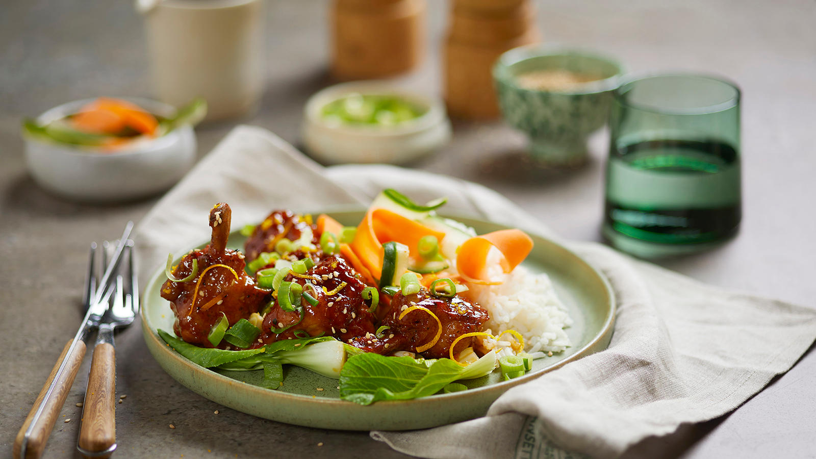 Kylling med asiatisk sticky glaze servert med bok choy, ris, gulrot, agurk, sitrusskall, vårløk og et dryss sesamfrø på lys grønn tallerken. Skåler med tilbehør i bakgrunnen.