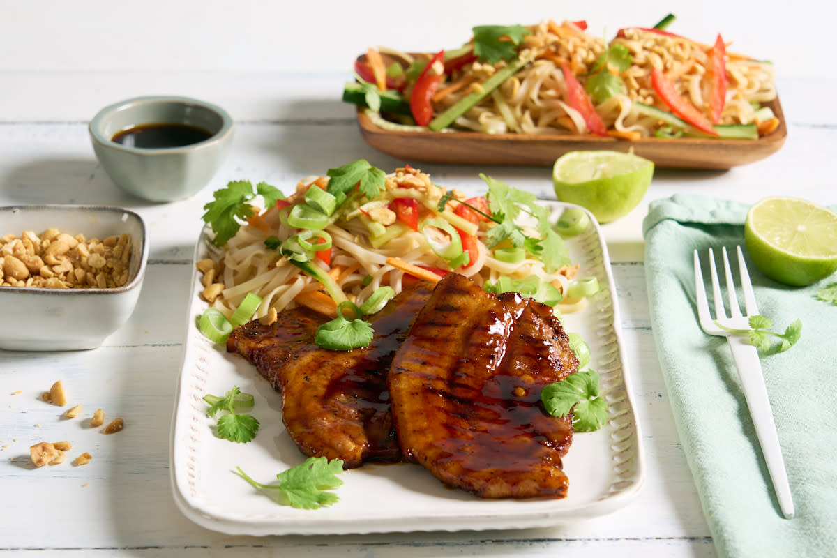 Asiatisk sticky grillribbe med kald nudelsalat servert på hvitt firkantet fat med limebåter, koriander og en skål peanøtter til. Firkantet trefat med kald nudelsalat i bakgrunnen.