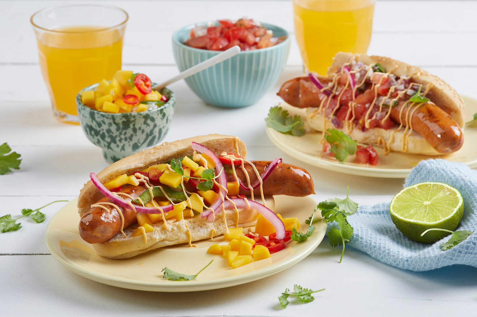 Cheesy Chili pølse med mangosalsa, pico de gallo og chilimajones servert i pølsebrød på lysegule tallerkner. Blå skåler i forskjellige mønster med tilbehør i bakgrunnen.