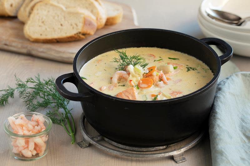 Kremet fiskesuppe med laks, torsk, reker og grønnsaker servert i sort støpejernskjele på lyst trebord. Skiver med loff, glass med reker, suppeskåler og linserviett på bordet rundt.