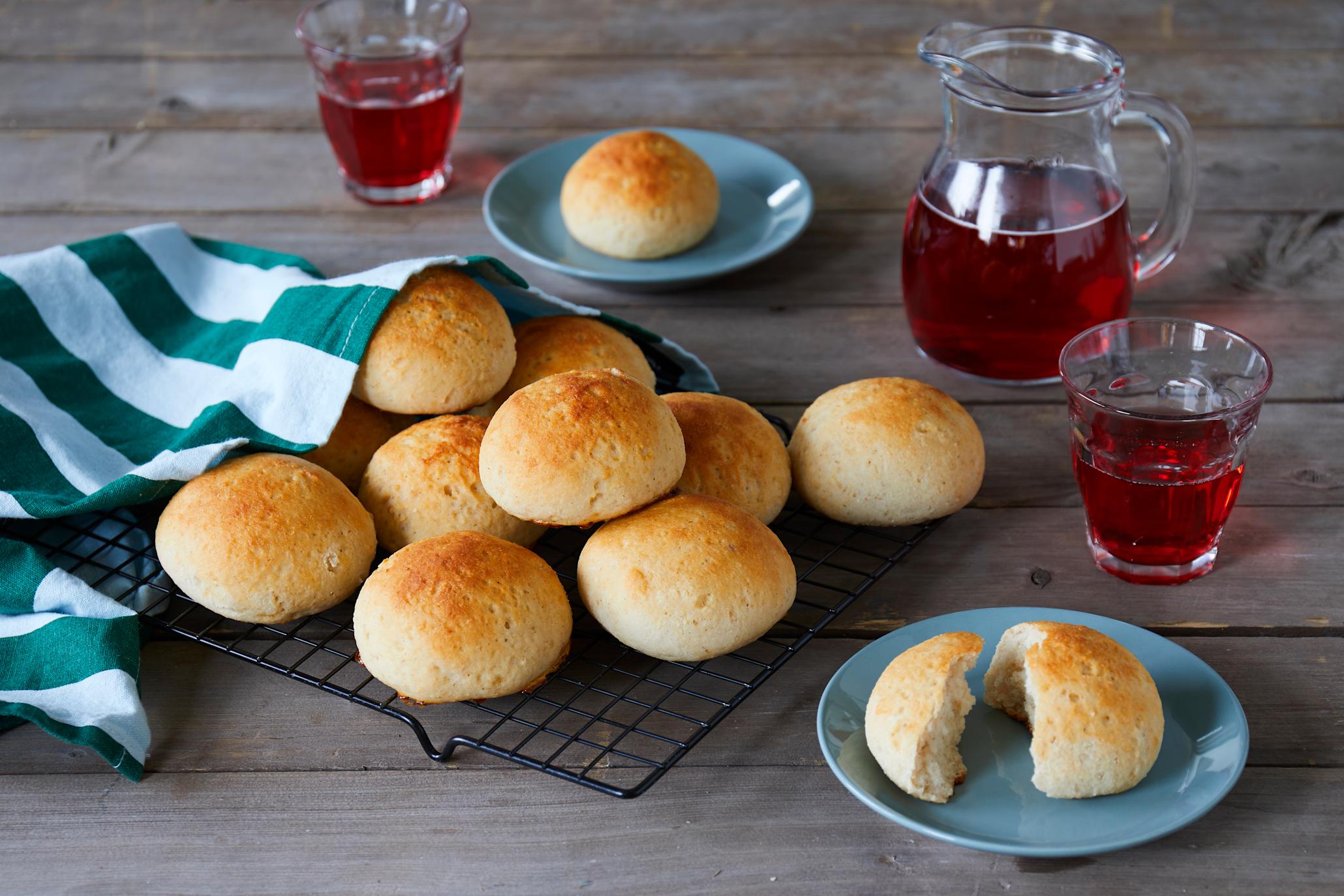 Glutenfrie boller på svart firkantet kjølerist, og servert på små turkise asjetter på rustikk trebord. Hvit og grønnstripete kjøkkenhåndkle ligger over bollene. Saftmugge og glass med rød saft i bakgrunnen.