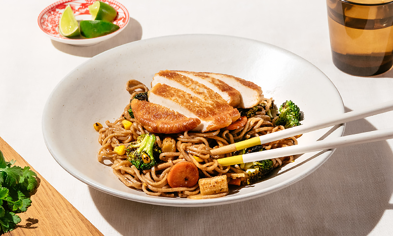 Fiskeburger med wok, nudler og grønnsaker. Servert i en skål med spisepinner, et glass vann og limebåter i bakgrunnen 