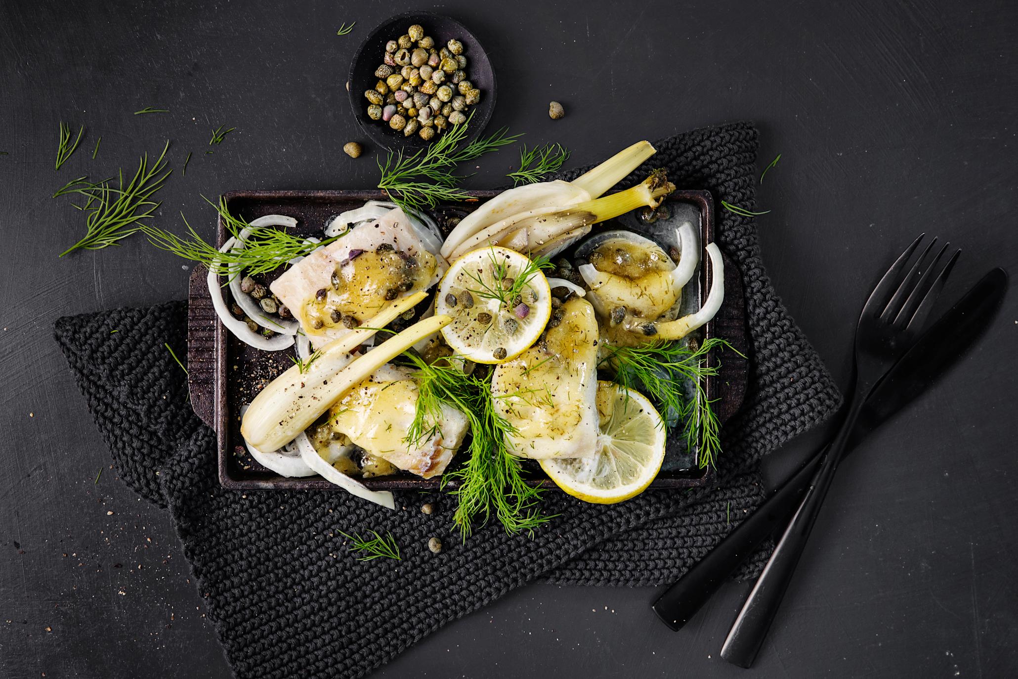Torsk i form med fennikel, kapers og sennepssaus fordelt utover svart ildfast form med frisk dill på toppen, på svart bord. Liten skål med kapers på siden.
