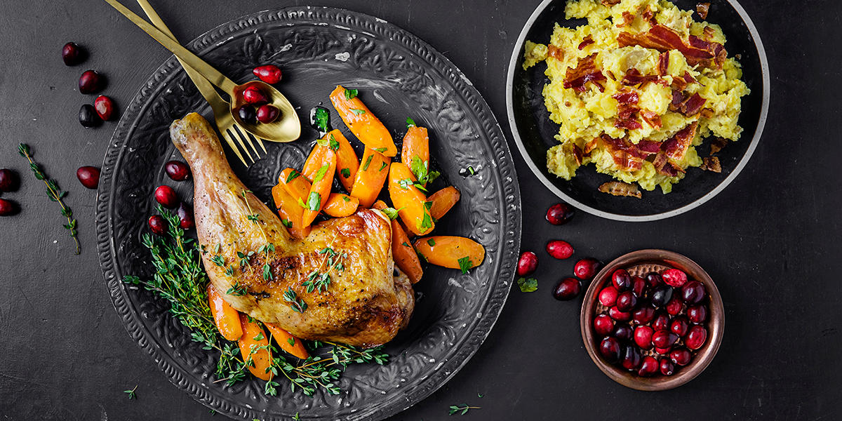 Ovnsbakt kalkunlår servert på mørkegrå tallerken, med braiserte gulrøtter med frisk persille. Mørk skål med potetmos med bacon drysset over på høyre side. Liten skål med tranebær ved siden av.