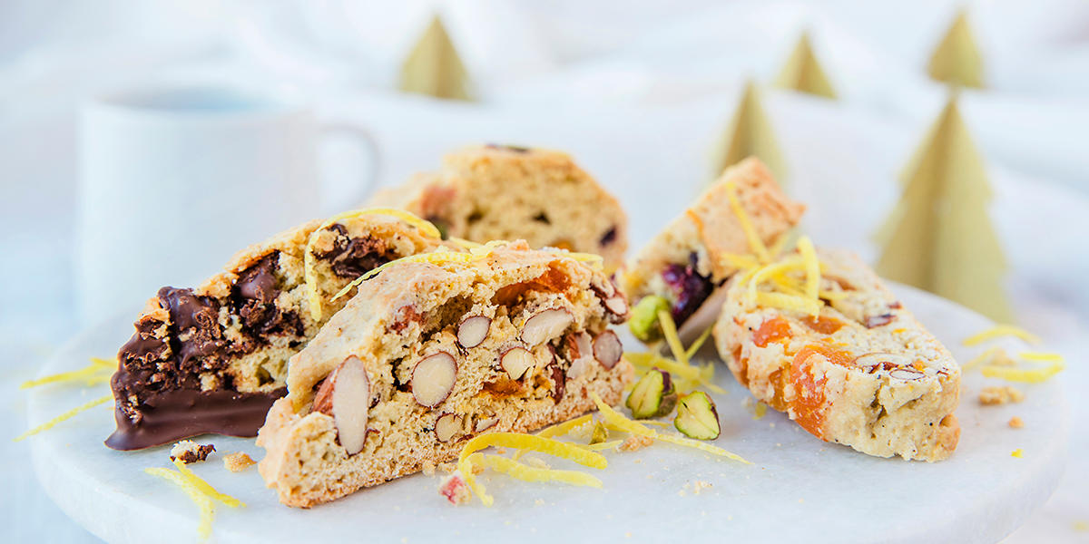 Biscotti på tre måter, servert på hvitt rundt marmorfat med hvit kaffekopp og gullfargede juletrær i papir i bakgrunnen. Biscottien har biter av nøtter, tørket frukt og sjokolade, og er drysset med revet sitronskall.