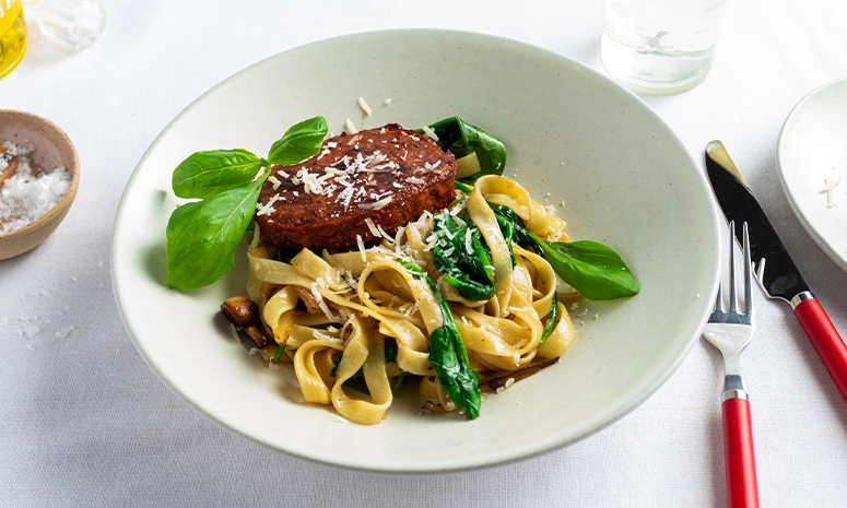 Vegansk biff, spinat og pasta i hvit skål pyntet med basilikum og parmesan.