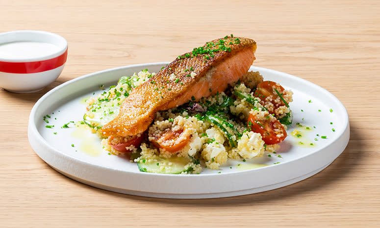 Ørret med couscous servert på en hvit tallerken, med høy kant, og en liten skål med yoghurt til venstre for tallerkenen.