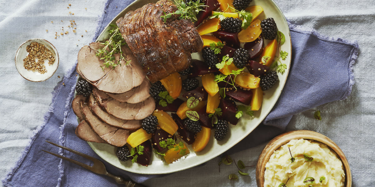 Rosastekt reinsdyrstek med glaserte beter servert på ovalt hvitt fat med ferske bjørnebær og urter. Fatet står på lys linduk med en treskål hjemmelaget potetmos ved siden av.