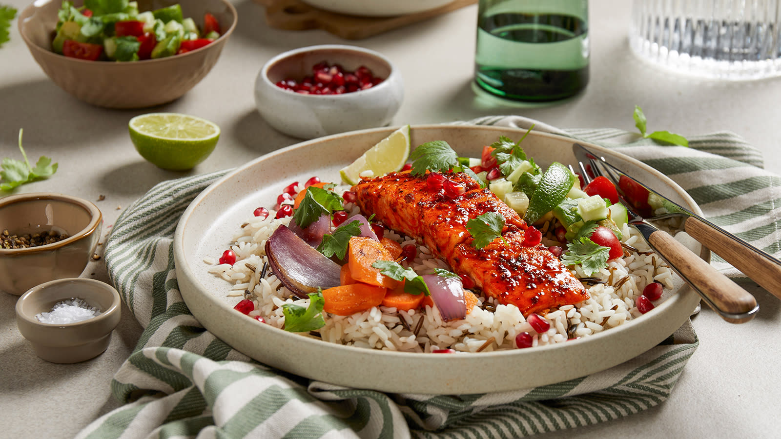 Stekt laks med stekte gulrøtter og avokadosalat servert på en seng av villris, og pyntet med friske urter og granateplekjerner. Skål med avokadosalat i bakgrunnen.